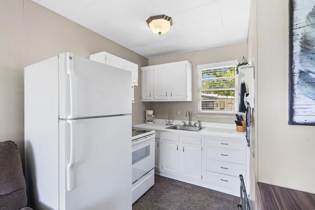 Compact cabin kitchen with white appliances, cabinets, and a window over the sink