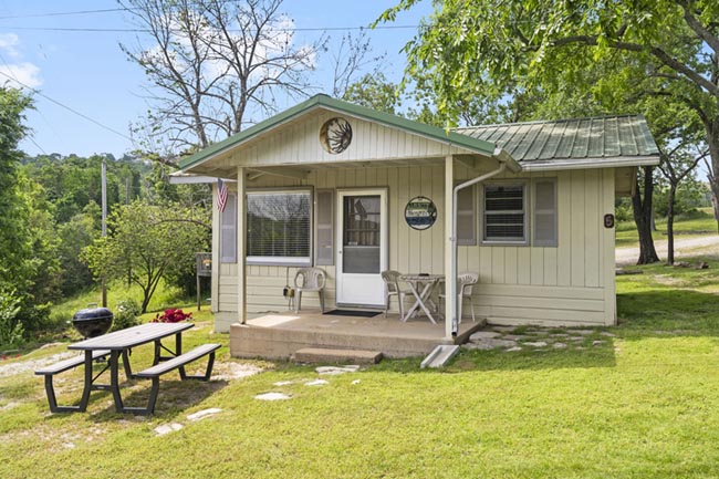Small beige cabin with a covered porch and picnic table on a grassy lakeside lot